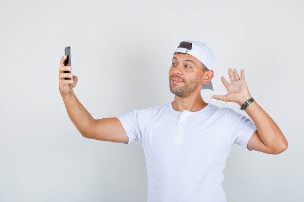 Jovem macho acenando com a mão enquanto tinha videocall em t-shirt branca, boné e olhando feliz, vista frontal.