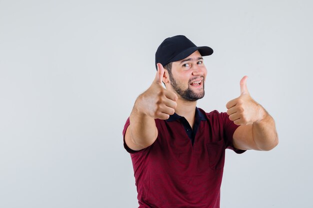 Jovem macho aparecendo o polegar em uma camiseta vermelha, boné preto e olhando feliz, vista frontal.