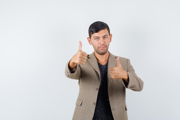 Jovem macho aparecendo o polegar em uma jaqueta marrom acinzentada, camisa preta e parecendo satisfeito. vista frontal.