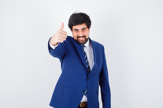 Jovem macho aparecendo o polegar na camisa, jaqueta, gravata e olhando alegre, vista frontal.