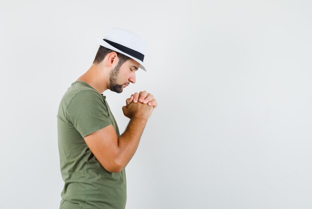 Jovem macho apertando as mãos em gesto de presa em t-shirt verde e chapéu e olhando esperançoso. .