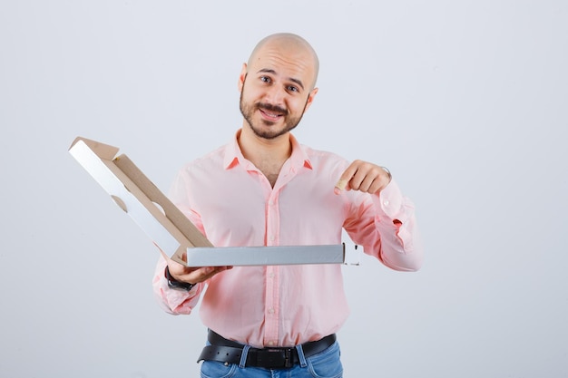 Jovem macho apontando abriu a caixa de pizza em camiseta, jeans e parece feliz. vista frontal.