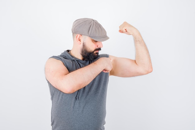 Jovem macho apontando os músculos do braço em um moletom sem mangas, boné e parecendo confiante. vista frontal.