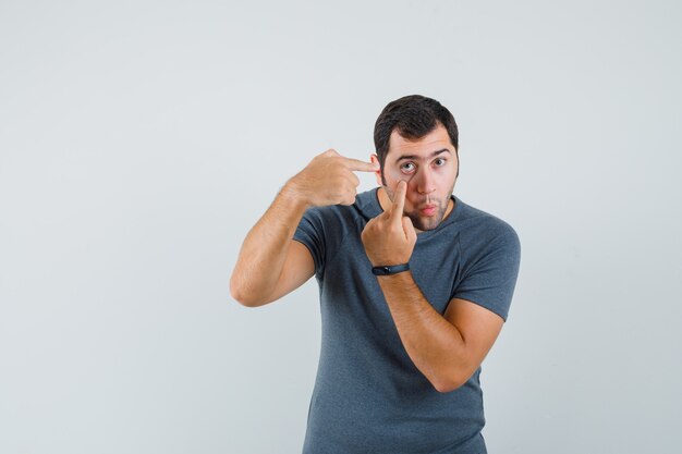 Jovem macho apontando para a pálpebra puxada pelo dedo em uma camiseta cinza