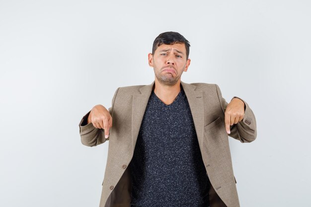 Jovem macho apontando para baixo em uma jaqueta marrom acinzentada, camisa preta e parecendo chateado. vista frontal.