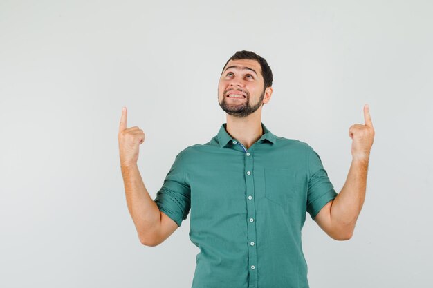 Jovem macho apontando para cima em camisa verde e olhando alegre, vista frontal.