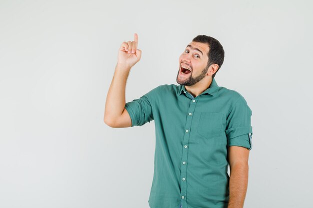 Jovem macho apontando para cima em camisa verde e olhando alegre, vista frontal.