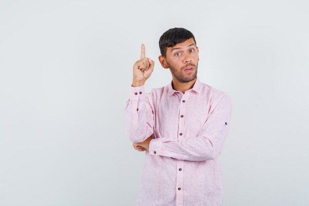 Jovem macho apontando para cima em uma camisa rosa e olhando cuidadoso, vista frontal.