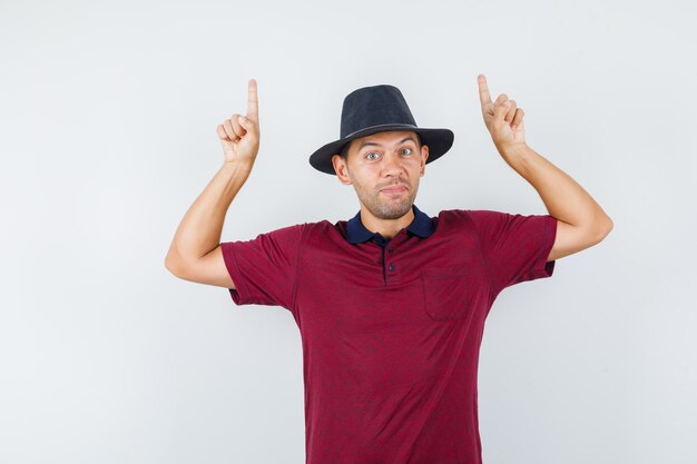 Jovem macho apontando para cima na camisa vermelha, vista frontal do chapéu preto.