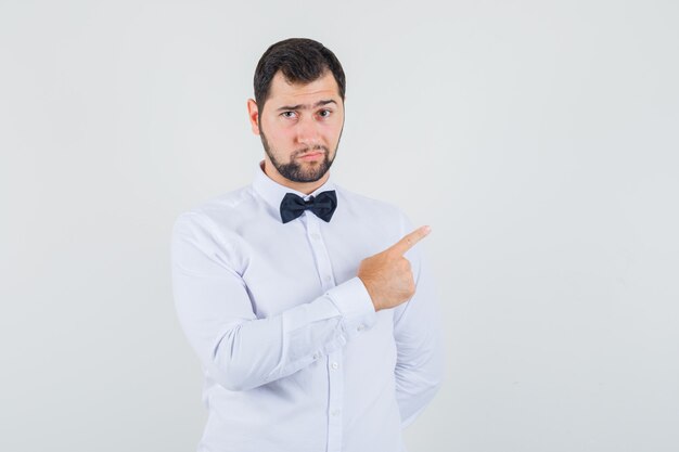 Jovem macho apontando para longe enquanto franzia a testa em uma camisa branca e parecendo confiante, vista frontal.