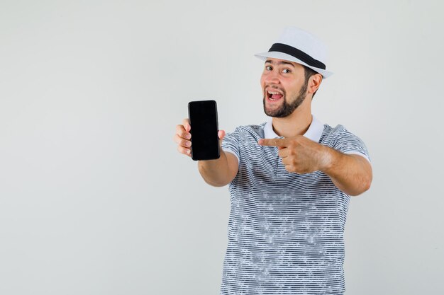 Jovem macho apontando para o celular em t-shirt, chapéu e olhando feliz, vista frontal.