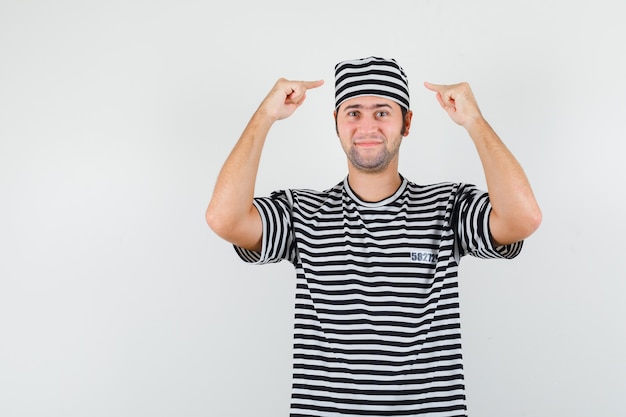 Jovem macho apontando para o chapéu em t-shirt, chapéu e olhando confiante, vista frontal.