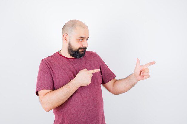 Jovem macho apontando para o lado em uma camiseta e parecendo confiante. vista frontal.