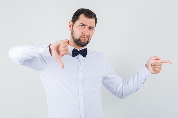 Jovem macho apontando para o lado, mostrando o polegar para baixo em uma camisa branca e parecendo descontente. vista frontal.