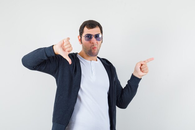 Jovem macho apontando para o lado, mostrando o polegar para baixo em uma camiseta, jaqueta e parecendo hesitante, vista frontal.