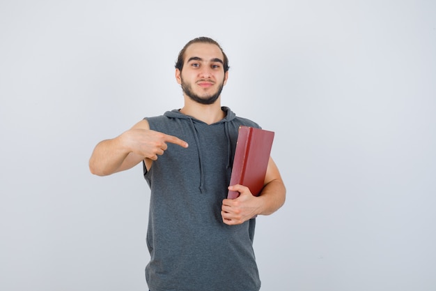 Jovem macho apontando para o livro com capuz sem mangas e olhando confiante, vista frontal.