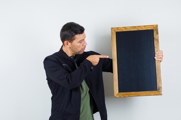 Jovem macho apontando para o quadro-negro em t-shirt, jaqueta e olhando focado, vista frontal.