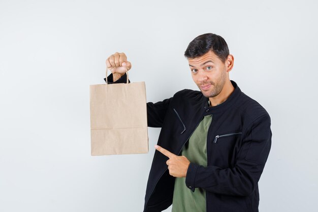 Jovem macho apontando para o saco de papel em t-shirt, jaqueta e olhando alegre, vista frontal.