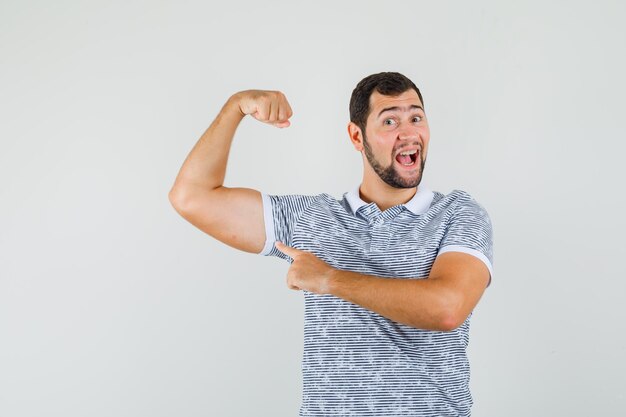 Jovem macho apontando para os músculos do braço em t-shirt e olhando alegre, vista frontal.