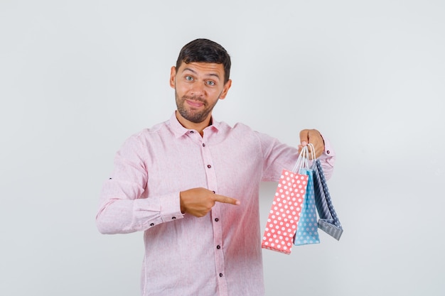 Foto grátis jovem macho apontando para sacos de papel na camisa e parecendo feliz. vista frontal.