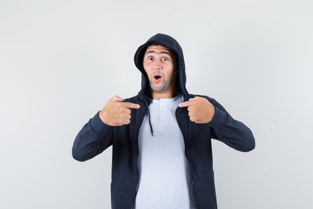 Jovem macho apontando para si mesmo em t-shirt, jaqueta e parecendo surpreso. vista frontal.