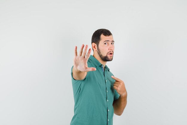 Jovem macho apontando para si mesmo enquanto mostra o gesto de parada na camisa verde e parecendo descontente. vista frontal.