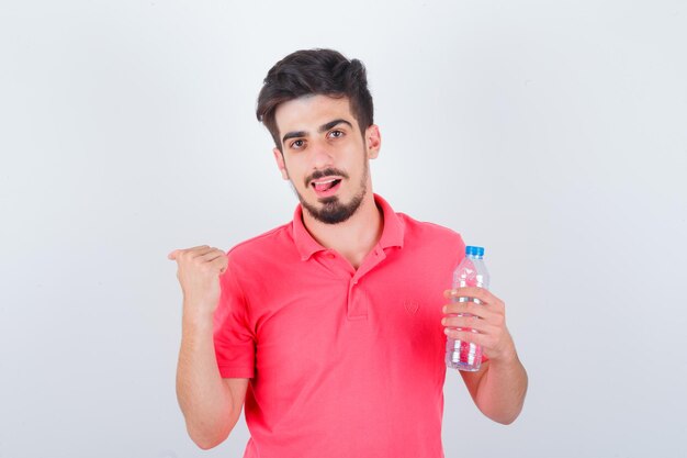 Jovem macho apontando para trás com o polegar enquanto mostra a língua na camiseta e parece feliz, vista frontal.