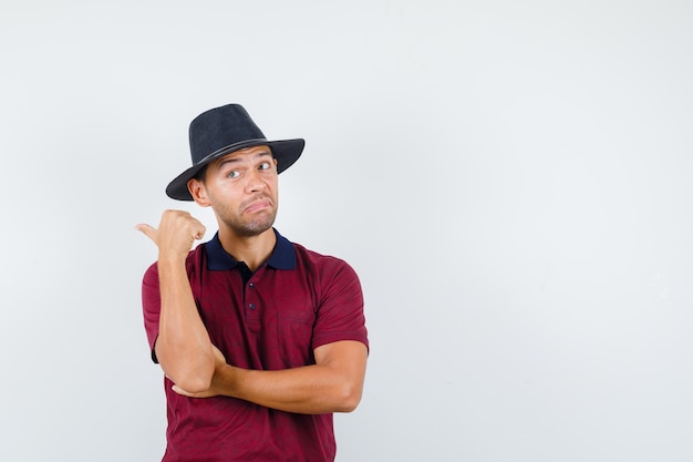 Jovem macho apontando para trás em camisa vermelha, chapéu preto e parecendo confuso, vista frontal. espaço para texto