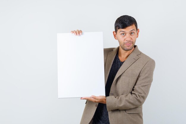 Jovem macho apontando para um papel em branco em uma jaqueta marrom acinzentada e olhando inteligente, vista frontal.