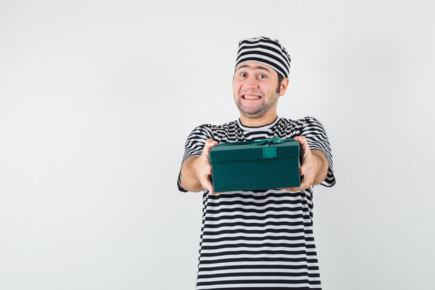 Jovem macho apresentando caixa de presente em t-shirt, chapéu e parecendo feliz. vista frontal.