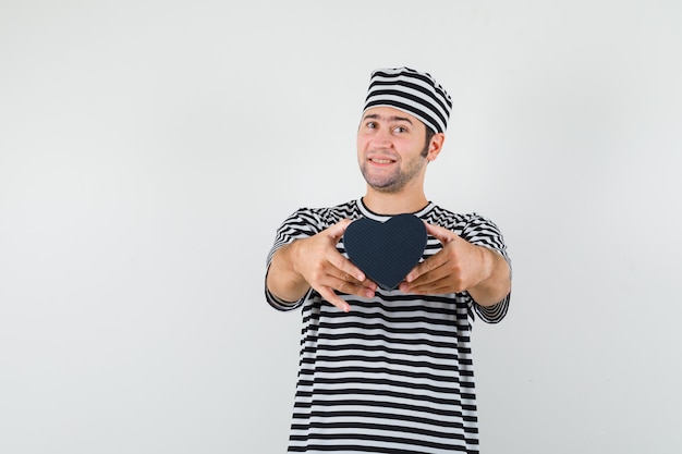 Jovem macho apresentando mini caixa de presente em camiseta listrada, boné e parecendo feliz. vista frontal. espaço para texto