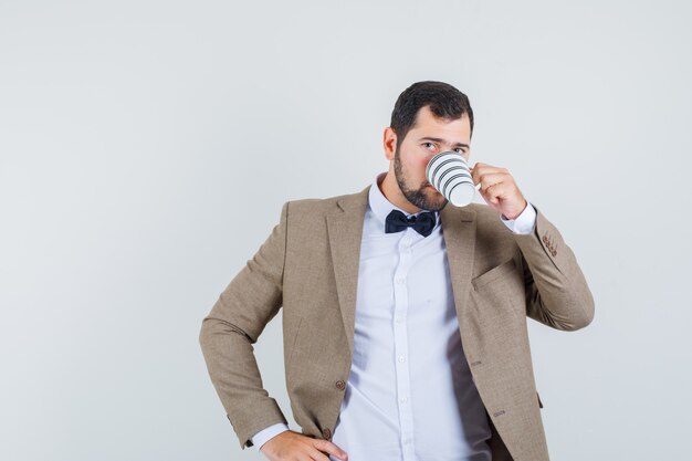 Jovem macho bebendo café com a mão na cintura no terno, vista frontal.