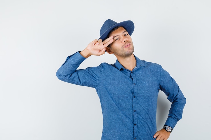 Foto grátis jovem macho com camisa azul, chapéu mantendo dois dedos perto das têmporas e olhando legal, vista frontal.