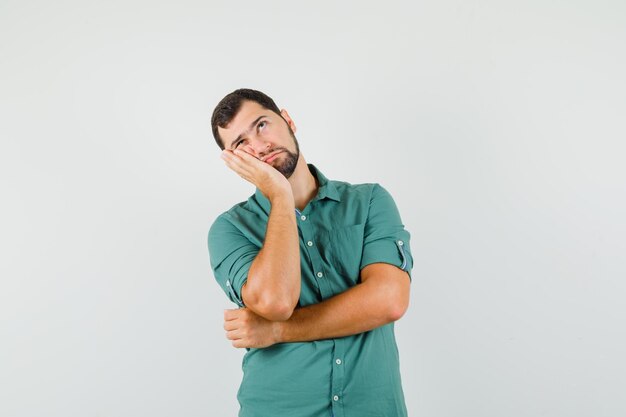 Jovem macho com camisa verde em pé e olhando pensativo, vista frontal.