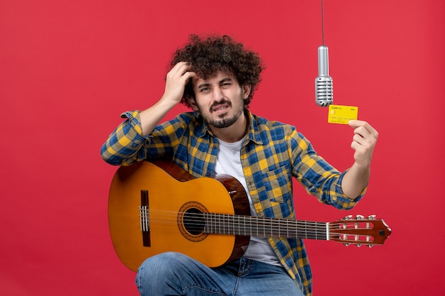 Jovem macho com guitarra, segurando o cartão do banco em uma parede vermelha.