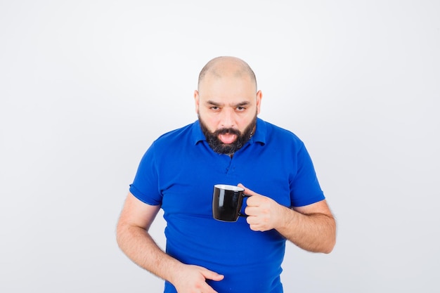 Jovem macho de camisa azul, segurando o copo enquanto coloca a língua para fora e olhando enojado, vista frontal.