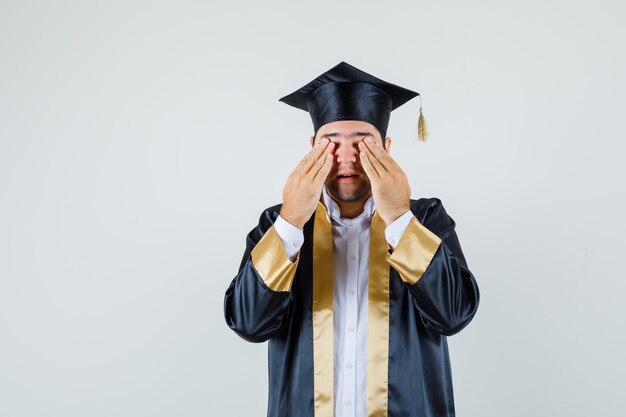 Jovem macho de mãos dadas nos olhos em uniforme de pós-graduação e parecendo animado. vista frontal.