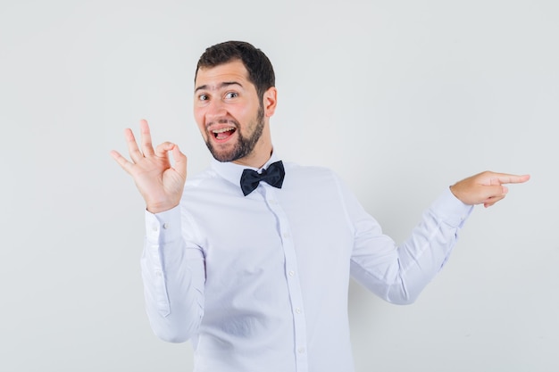 Jovem macho em camisa branca, apontando para o lado, mostrando o gesto de ok e olhando alegre, vista frontal.