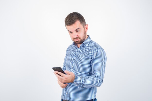 Jovem macho em camisa, jeans, olhando para o celular e parecendo perplexo, vista frontal.