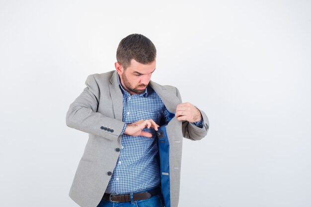 Jovem macho em camisa, jeans, paletó, olhando para o bolso da jaqueta e olhando sério, vista frontal.