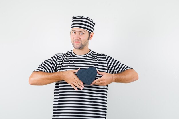 Foto grátis jovem macho em camiseta listrada, boné segurando mini caixa de presente, vista frontal.