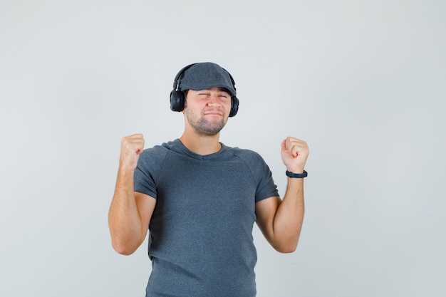 Jovem macho em t-shirt, boné curtindo música com fones de ouvido e olhando feliz, vista frontal.