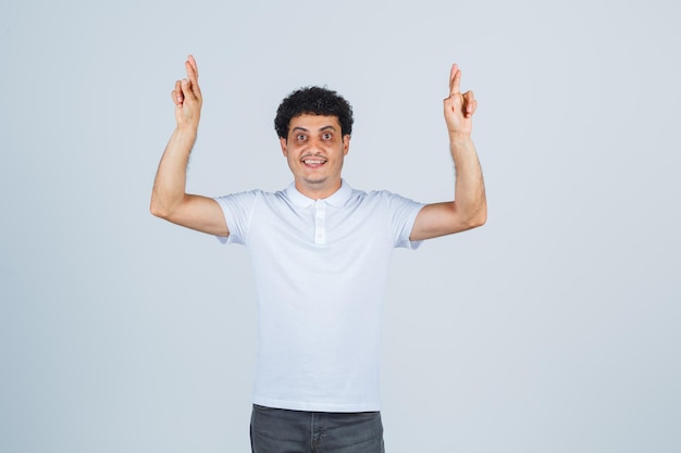 Jovem macho em t-shirt branca, calça mantendo os dedos cruzados e parecendo feliz, vista frontal.