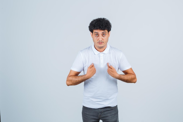 Jovem macho em t-shirt branca, calça mostrando-se e parecendo surpreso, vista frontal.