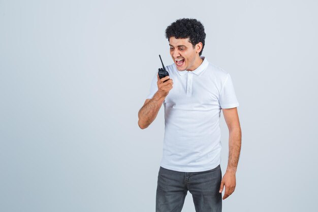 Jovem macho em t-shirt branca, calças usando o telefone walkie-talkie da polícia e olhando animado, vista frontal.