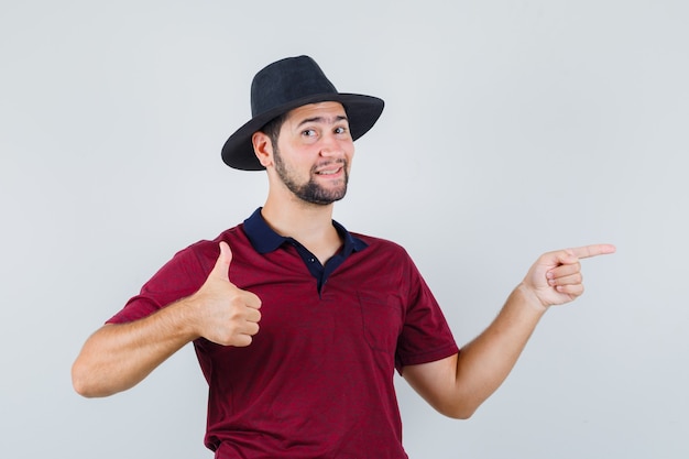 Jovem macho em t-shirt, chapéu apontando para o lado enquanto aparecia o polegar e parecendo satisfeito, vista frontal.