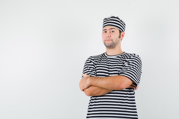 Jovem macho em t-shirt, chapéu em pé com os braços cruzados e olhando confiante, vista frontal.