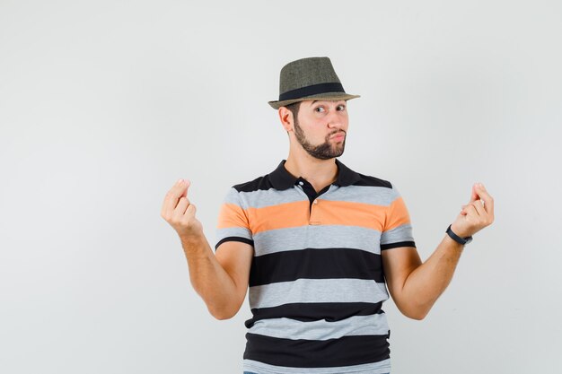 Jovem macho em t-shirt, chapéu gesticulando com as mãos e dedos e olhando confiante, vista frontal.