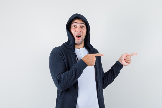 Jovem macho em t-shirt, jaqueta apontando para o lado e parecendo espantado, vista frontal.