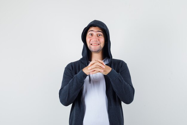 Jovem macho em t-shirt, jaqueta, mantendo os dedos entrelaçados e olhando alegre, vista frontal.